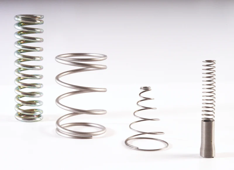 Cuatro tipos de muelles metálicos industriales sobre fondo blanco: un muelle de compresión largo, un muelle helicoidal ancho, un muelle cónico y un muelle fino con base reforzada.