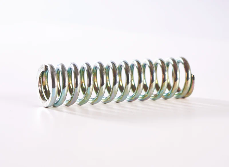 Primer plano de un muelle de compresión de acero grueso dispuesto horizontalmente sobre un fondo blanco, mostrando un acabado metálico brillante y espiras uniformes de alta resistencia.