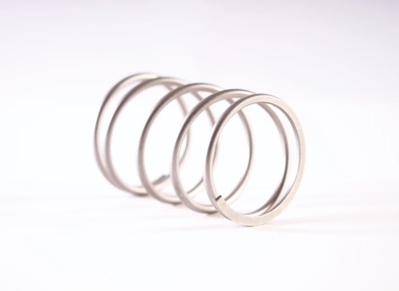 Primer plano de un muelle de compresión de acero inoxidable con espiras de gran diámetro y acabado mate, orientado diagonalmente sobre un fondo blanco neutro.