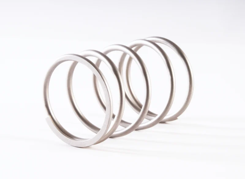 Vista en perspectiva de un muelle helicoidal de acero inoxidable con espiras anchas y regulares, resaltando el acabado metálico satinado sobre un fondo blanco limpio.