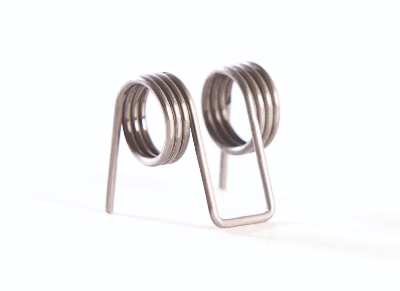 Muelle de torsión doble de acero sobre fondo blanco, con dos bobinas helicoidales conectadas por un puente central en forma de U y brazos de apoyo laterales.