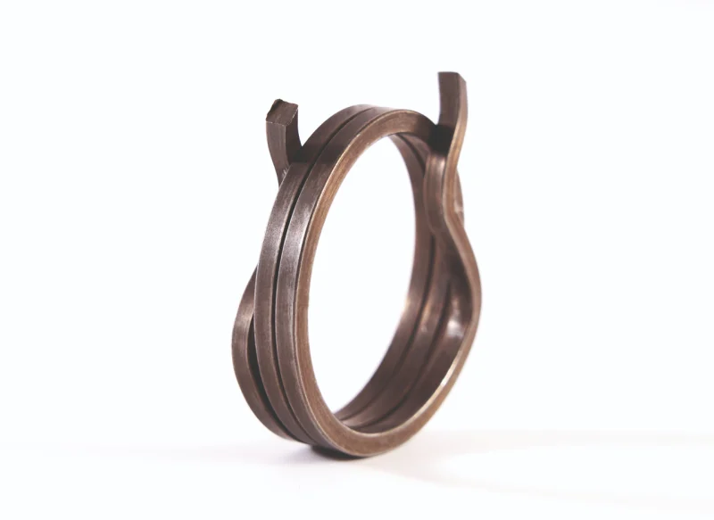 Muelle de torsión de acero sobre fondo blanco, con un cuerpo de espiras juntas y un brazo metálico largo que termina en un gancho en forma de U para sujeción mecánica.